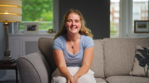 Libby sitting on a sofa, smiling at the camera, wearing a pale blue t-shirt and white skirt.