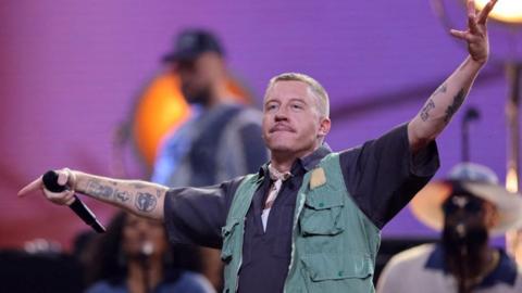 Macklemore on stage, his arms thrown in the air. He is holding a microphone in his right hand and is looking out at the crowd, with a band behind him