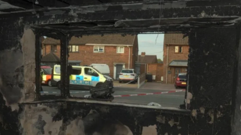 A police van is visible on the street through the burnt out windows of the house. There is no glass in the frame and police tape outside the property is also visible.