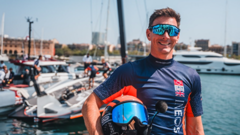 A man in a navy wet suit and sun glasses holding a helmet in a harbour