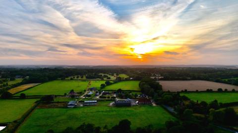 A setting sun shines through wispy clouds as the sky behind them graduates from orange to blue. The lower half of the picture is a green landscape of fields and trees with a farm and stables