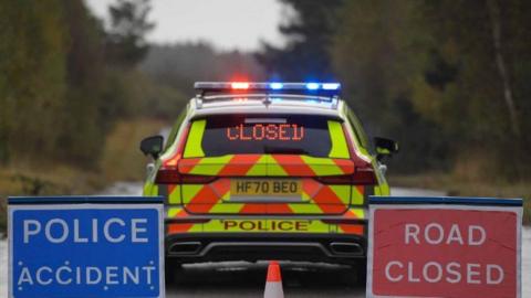 Lights in rear windscreen of police car spell closed with red road closure sign on right and blue police accident sign on left