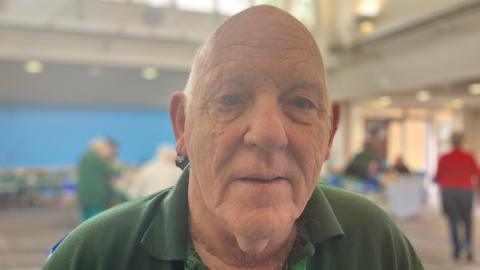 A head-and-shoulders photo of a bald man wearing a green polo shirt. In soft focus behind him there's a church hall with people milling around.