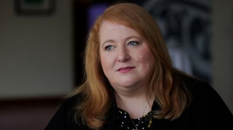 A woman with red shoulder-length hair looks off to the side of the camera. She's wearing a patterned top with a black jacket.