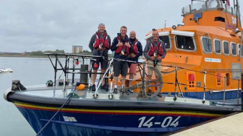 Four RNLI crew stand on the deck of an orange and blue all-weather Trent Class lifeboat.