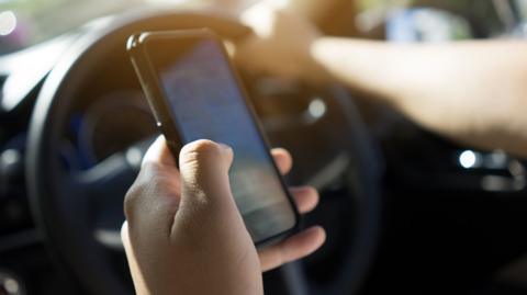 A hand holding a phone next to a steering wheel. The person's other hand is on the wheel.