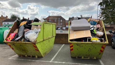 Two lime green skips full of rubbish in a car park side by side. One has a mattress and a large piece of carboard, as well as a bike frame, while another has plenty of bags full of rubbish