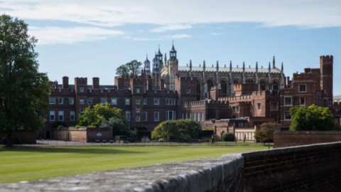 Eton College seen from a distance. It's a large red brick building with multiple spires and turrets, with a well-kept lawn next to it.