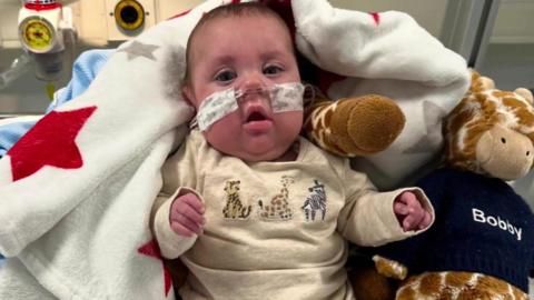 A baby with a tube in his nose. He is laying next to a cuddly toy giraffe.