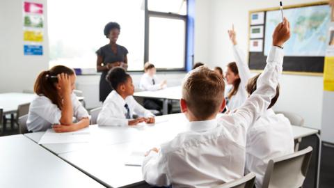 Students are sat in a classroom, wearing white shirts and ties, some have their hands raised to answer a question set by the teacher standing in the classroom 