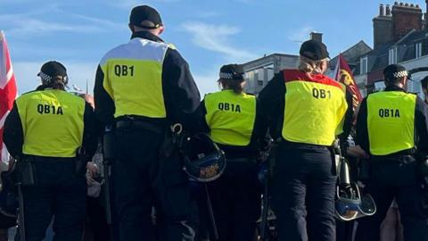 Rear view of a line of police officers standing in front of a crowd waving Union flags and banners. In the foreground is a dog handler and a dog