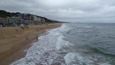 Boscombe beach on an overcast day. There are a few people on the beach with some children paddling. There are waves breaking onto the beach with white foam and the sky overhead is grey. Behind the beach there are several buildings in front of the cliff and there is a lifeguard station on the beach. 