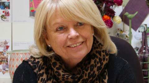 Head shot of Gisela Graham who has blond shoulder length hair and is wearing a black top and leopard skin style silk scarf