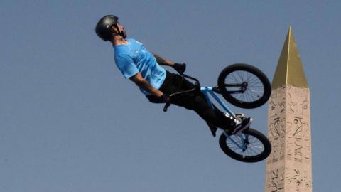 The camera angle creates the appearance that a BMX rider is riding up the side of an obelisk at the Paris Olympics