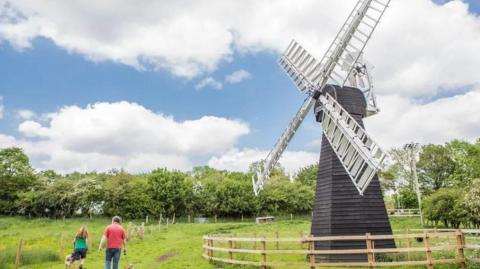 Two people are walking past a large black windmill with white sails at the museum