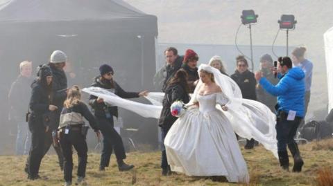 Margot Robbie wearing a voluminous wedding dress and veil and carrying a bouquet of flowers. She is surrounded by film crew and cameras. 