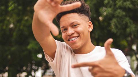 A teenage boy outdoors smiles at the camera and frames himself in the shot with his hands.