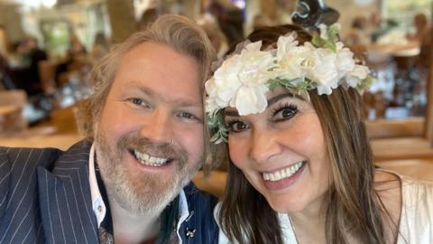 Keaton and Sophia Stone smile at the camera with their faces close together. He is wearing a pinstripe suit and she is wearing a white dress and a white flower crown on her head. He has light grey hair and a beard, while she has dark brown hair.