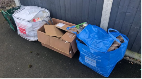 Three bags left on the side of the pavement. One is a white bag, a cardboard box and a blue bag. They are all used for waste and recycling