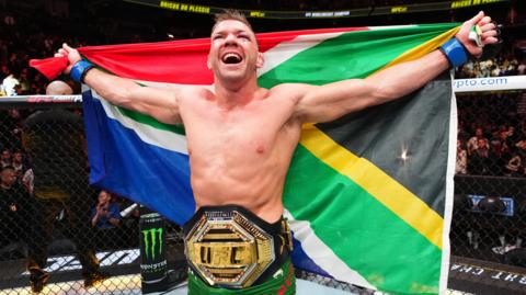 Dricus du Plessis celebrates in the Octagon with a UFC title belt around his waist and South African flag held up behind him