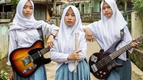 Three women wearing hijabs standing together. Two are holding guitars and one is holding drum sticks.
