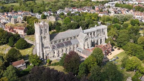 An aerial view of Christchurch Priory. The large grey stone building has a large tower at one end with a clock. Behind you can see the ruins of Christchurch Castle and the homes and shops of the town. The area of is full of lush green trees. 