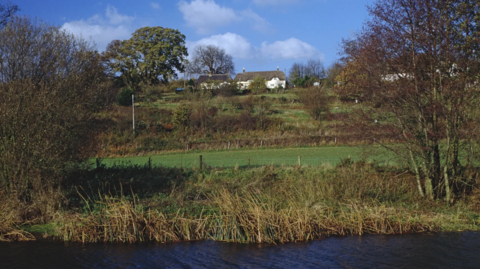 A murky river with reeds. On the river bank is a wire fence and trees. Fields rise up a small hill to a handful of white properties at the top.