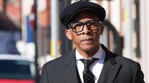 Image shows Jay Blades arriving at Worcester Crown Court wearing a black suit, white shirt, black tie with white spots and a tie pin and a red handkerchief tucked into his breast pocket. He is wearing a black peaked cap.