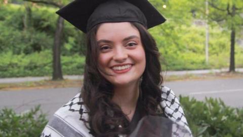 Aysenur Ezgi Eygi smiles and looks at the camera. She has dark hair and is wearing a mortar board hat, showing that she is possibly graduating, and has a keffiyeh draped around her shoulders 