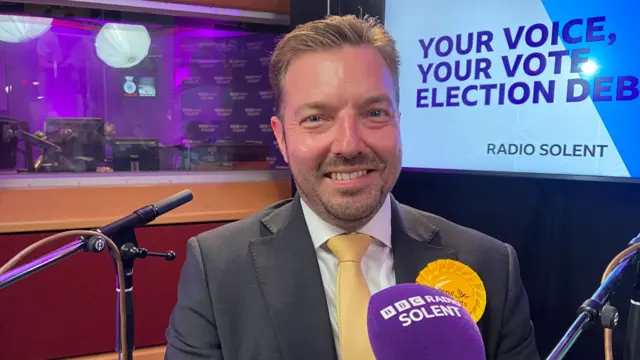 Geoff Cooper in a green suit with yellow tie, sitting in front of a microphone