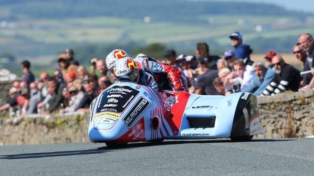 Ben Birchall and Kevin Rousseau racing in a light blue and red sidecar outfit with black sponsorship panels. It has the number 16 on the front in white and they are wearing leathers that match the outfit but have yellow three-legs-of-Mann symbols on the side. A stone wall curves round behind them with spectators watching from behind it.