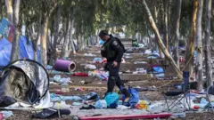 Policial caminhando pelo local onde foi realizado o festival de música, em meio a lixo, restos de barracas e colchonetes