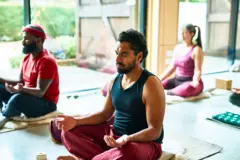 Foto colorida mostra três pessoas sentadas no chão com as pernas cruzadas e olhos fechados, em posição de meditação