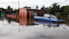 Carro em meio a água, com casas ao fundo