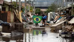 Uma mulher segura uma bandeira do Brasil em meio a diversos móveis em uma rua ainda com sinais de água em Porto Alegre