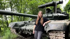 Jovem branca e loira em frente a um tanque de guerra