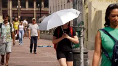 Pessoas caminham na rua durante onda de calor em São Paulo. Uma delas usa um guarda-chuva para se proteger do sol