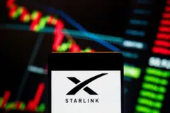 Logotipo da Starlink exibido em um smartphone com um gráfico de índice econômico da bolsa de valores ao fundo.