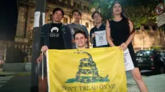 Jovens posando para foto à noite; um deles segurando uma Bandeira de Gadsden