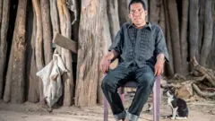 Chagabi Etacore sitting in a chair in the forest with a cat beside him