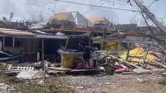 Mulher chora em frente  a destroços de uma casa e um poste caído