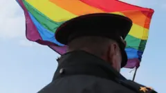 Um oficial russo em frente a uma bandeira do movimento LGBTQIA+