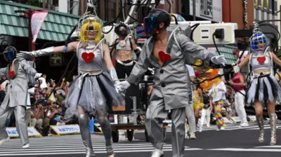 Desfile de uma escola de samba em uma rua. No primeiro plano, um homem e uma mulher de mãos dadas, fantasiados com uma roupa cinza e um coração vermelho no peito.