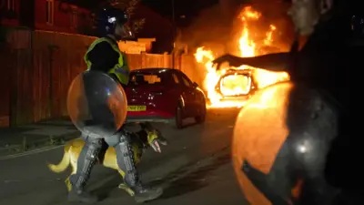 Policial passa com cão em rua com um carro em chamas ao fundo 