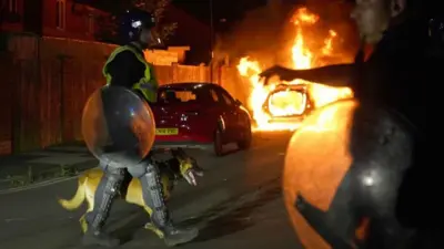 Policial com cachorro passa em frente a carro pegando fogo em protesto