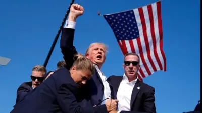 Trump com rosto ensanguentando - ao seu redor, agentes do Serviço Secreto. No fundo, uma bandeira dos EUA
