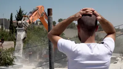 Ahmad stands with hands on head as a bulldozer demolishes his home