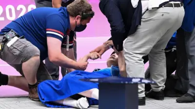 Atleta caída no chão com pessoas em volta tentando ajudar