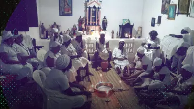 Pessoas de branco reunidas e sentadas em sala; há imagens de santos e velas no chão 