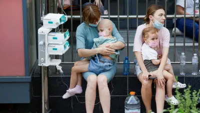 Crianças no colo de cuidadores do lado de fora, após a explosão no Hospital Infantil Ohmatdyt